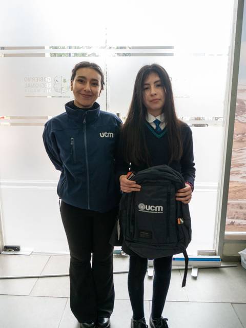 Dos personas posan juntas en un entorno interior, una de ellas sostiene una mochila.