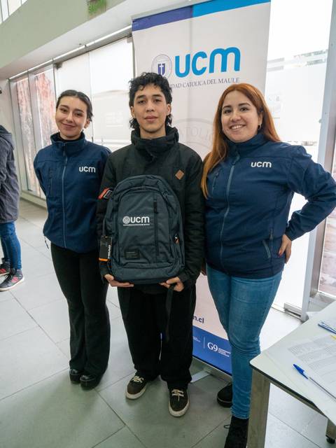 Tres personas posan juntas, dos mujeres a los lados y un hombre en el centro sosteniendo una mochila, frente a un banner de la UCM.