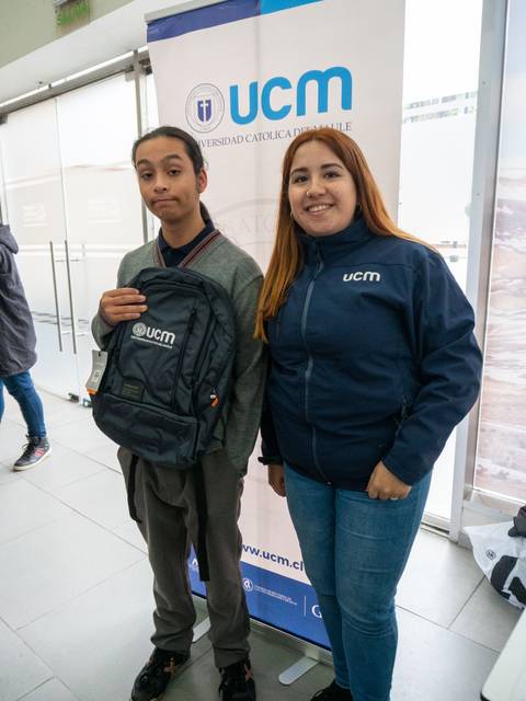 Dos personas posan frente a un banner de la Universidad Católica del Maule, una de ellas sostiene una mochila.