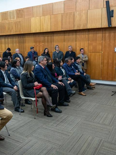 Un grupo de personas asiste a un evento en una sala con banderas chilenas en el fondo.