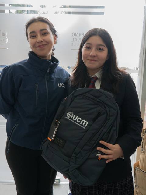 Dos jóvenes posan juntas, una de ellas lleva una mochila y ambas sonríen ante una puerta de vidrio.