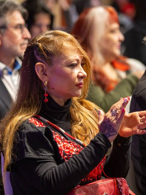 Una mujer con cabello largo y suelto, vestida con un top negro y un chal rojo, se encuentra entre un grupo de personas sentadas, aplaudiendo en un evento.