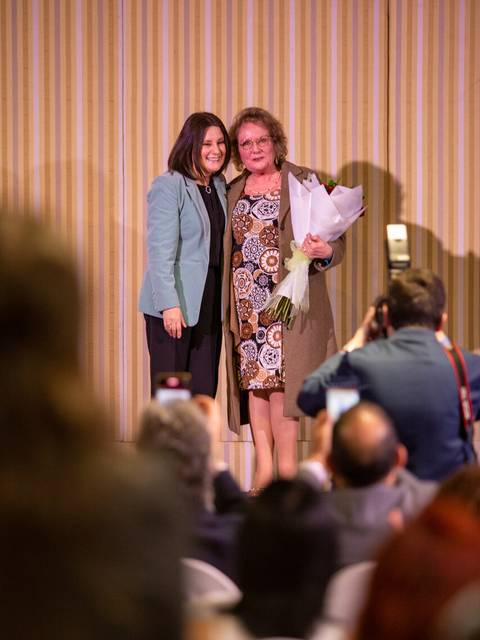 Dos mujeres sonrientes posan juntas en un evento formal, una de ellas sostiene un ramo de flores.