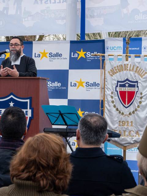 Un orador se presenta en un evento al aire libre frente a un público reunido en torno a un podio con el escudo de un colegio.