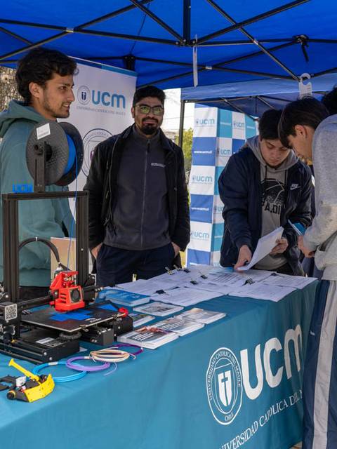 Un grupo de estudiantes están reunidos en un stand de la UCM, donde se exhiben impresoras 3D y materiales educativos.
