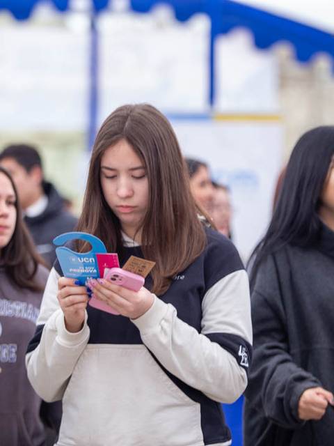 Un grupo de jóvenes en un evento al aire libre, con una chica en primer plano revisando documentos o tarjetas.