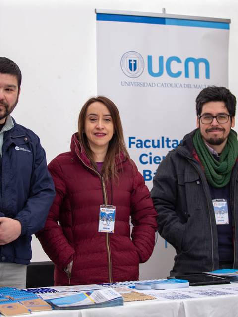 Tres personas posan frente a un stand de la Universidad Católica del Maule durante un evento.