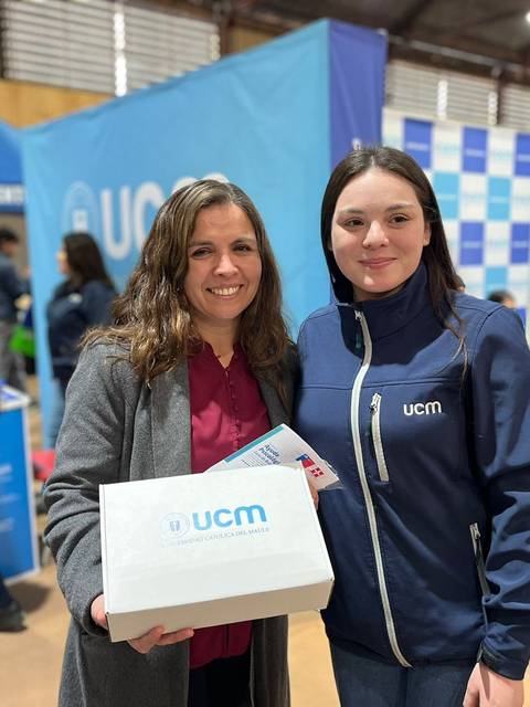 Dos mujeres sonríen frente a un evento con estands decorados, una de ellas sostiene una caja con el logo de UCM.
