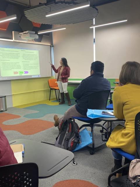 Una mujer está presentando en una sala de clases moderna mientras los asistentes están escuchando.