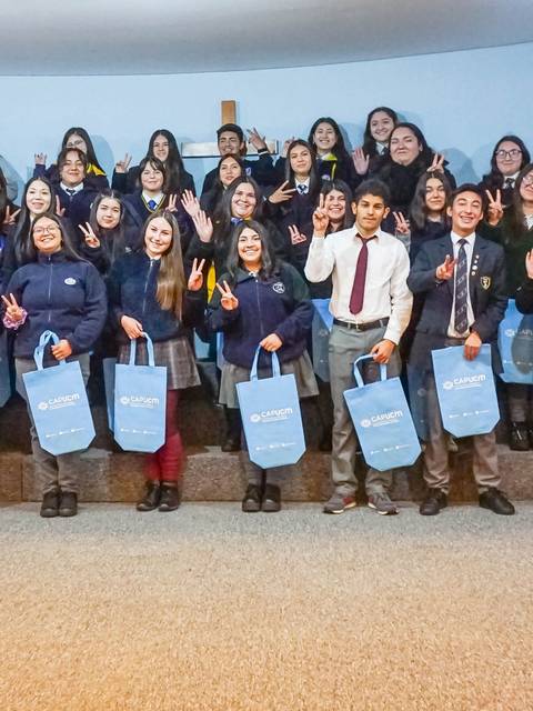 Un grupo numeroso de estudiantes sonríen y posan frente a la cámara, muchos de ellos levantando la mano en señal de saludo, mientras sostienen bolsas azules.
