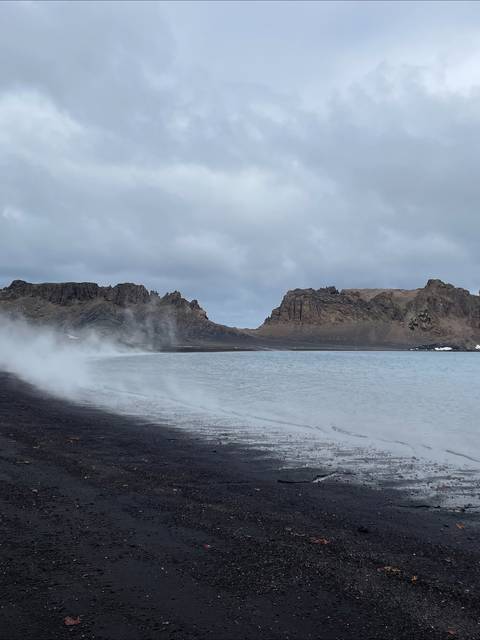 Una playa con arena negra y mar azul, rodeada de montañas y nubes grises.