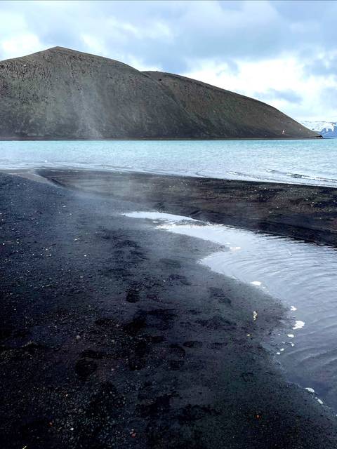 Una playa de arena negra con montañas al fondo y agua clara.