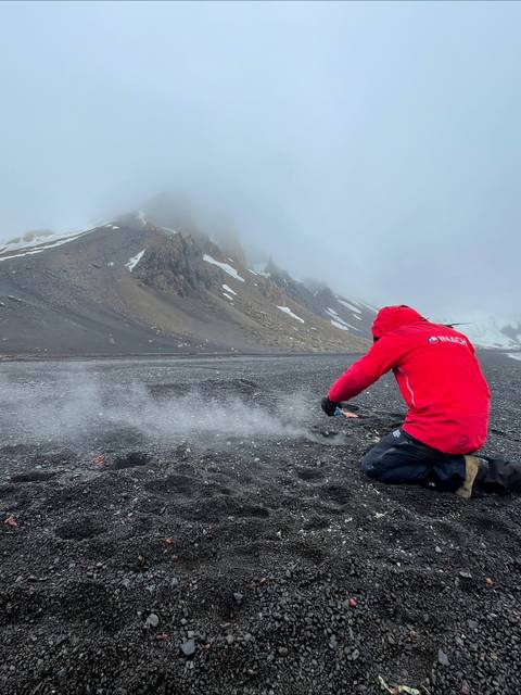 Una persona con chaqueta roja está agachada en una playa de arena oscura, observando el paisaje montañoso y nublado a su alrededor.
