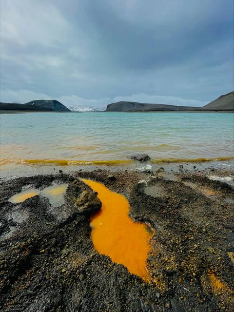 Una playa rocosa con agua de color naranja en un paisaje montañoso bajo un cielo nublado.
