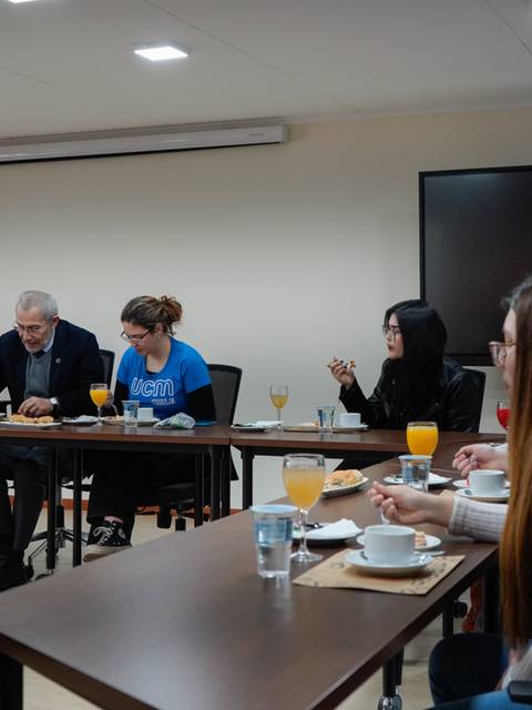 Un grupo de personas se encuentra sentado alrededor de una mesa en una reunión con alimentos y bebidas.