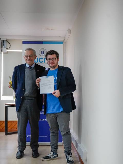 Dos hombres posan juntos, uno de ellos sosteniendo un certificado en un evento académico.