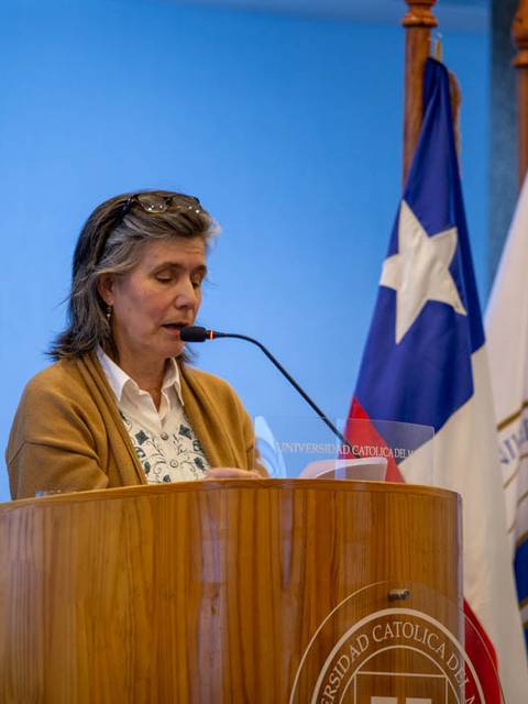 Una mujer habla en un podio frente a banderas de Chile y de una universidad católica.
