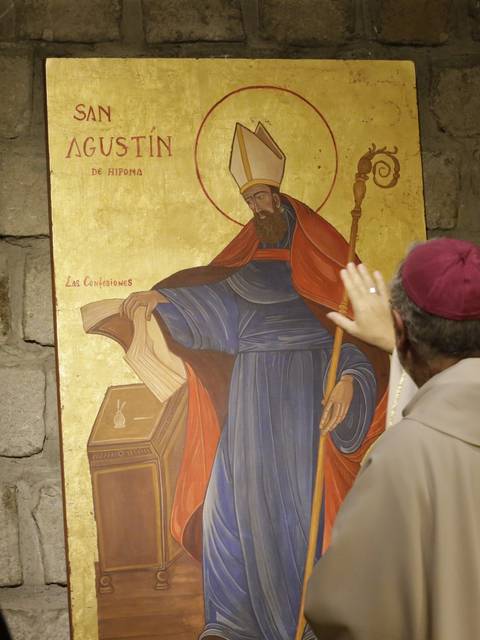 Un hombre de espaldas viste una vestimenta religiosa mientras señala una pintura de San Agustín en la pared.
