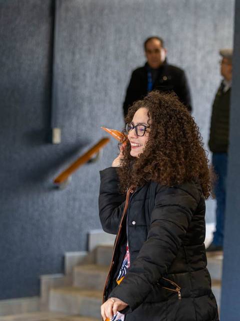 Una mujer con cabello rizado y gafas sonríe mientras sostiene un objeto en la mano en un entorno interior.