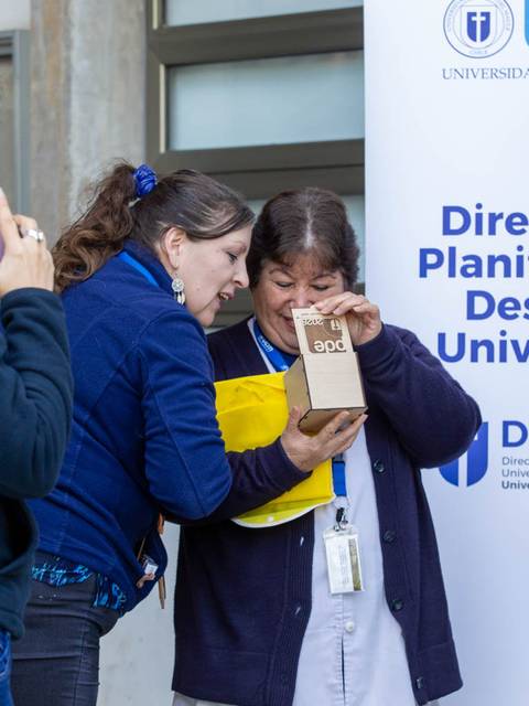 Dos mujeres están sonriendo y compartiendo un momento de sorpresa al abrir un paquete en un evento en la universidad.
