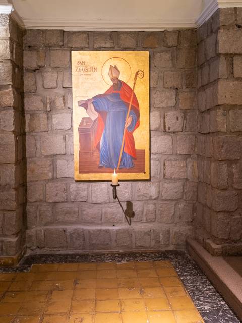 Un hombre de pie junto a una pintura religiosa en un interior de iglesia con paredes de piedra.