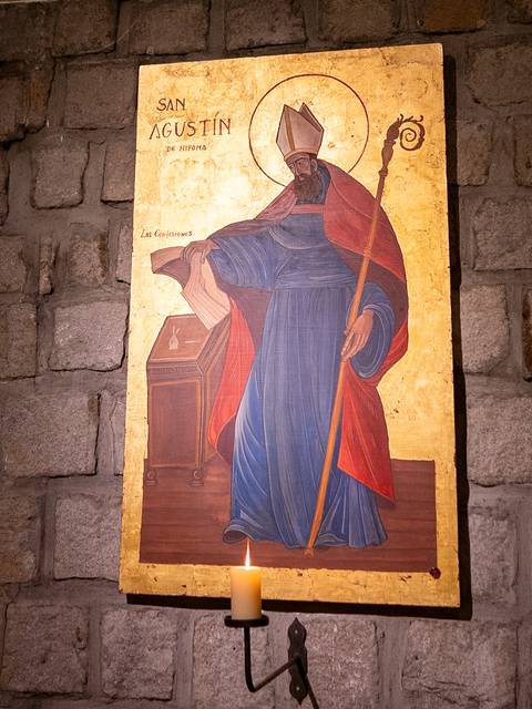 La imagen muestra una iconografía de San Agustín en un entorno con paredes de piedra.