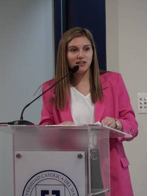 Una mujer de cabello largo y lacio, vestida con una chaqueta rosa, habla frente a un podio en un evento.