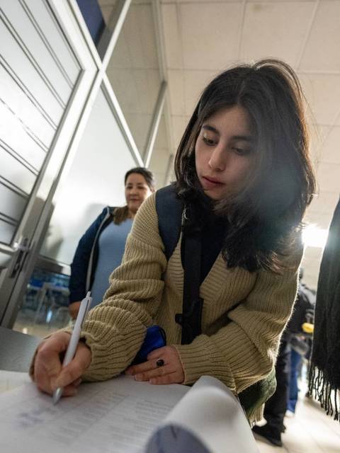 Una joven se encuentra firmando un documento en un entorno académico, mientras otra persona la observa al fondo.