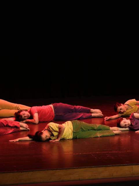 Grupo de bailarines en una coreografía en el escenario, vestidos con ropa de colores brillantes.