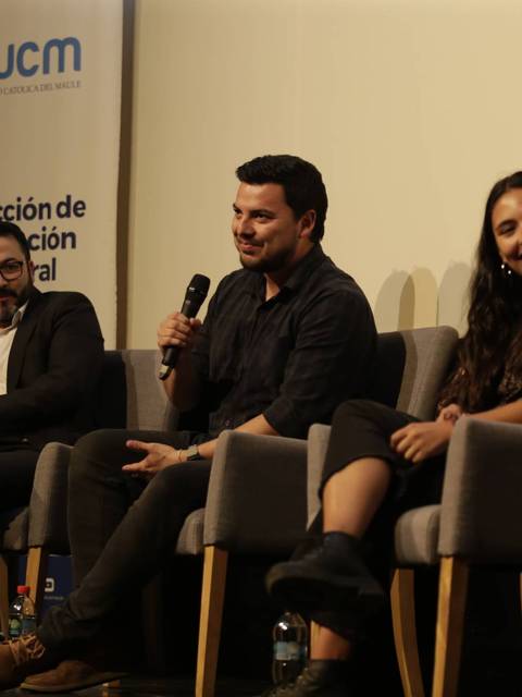 Un panel de discusión con cuatro personas sentadas en un escenario, con una bandera y un banner de la universidad al fondo.