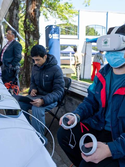 Una persona usa gafas de realidad virtual mientras otra está sentada revisando un dispositivo portátil en un evento al aire libre.