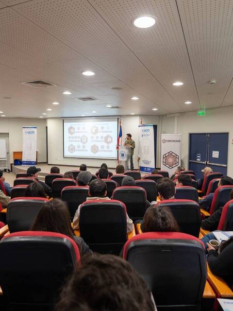 Una conferencia en un salón con asistentes sentados y un ponente en el escenario.