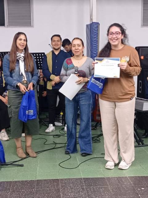 Un grupo de personas en un evento de premiación, posando sonrientes con diplomas y bolsas, mientras otros músicos están al fondo.