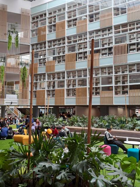 Un espacio interior moderno con plantas, áreas de descanso y grupos de personas conversando.