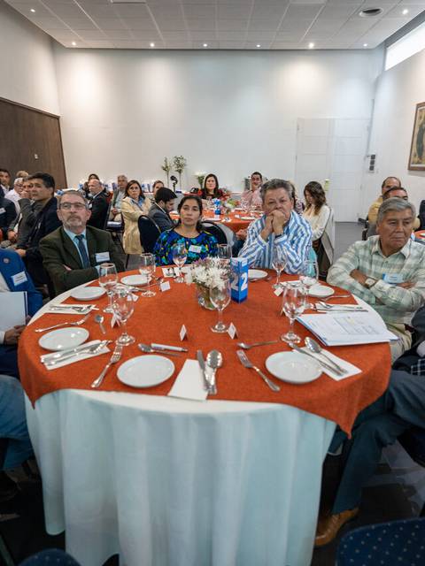 Un grupo de personas se reúne en una sala para un evento formal sentado alrededor de mesas con manteles naranjas.