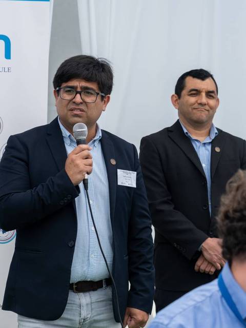 Dos hombres en un evento académico, uno hablando por un micrófono frente a una presentación de la universidad.