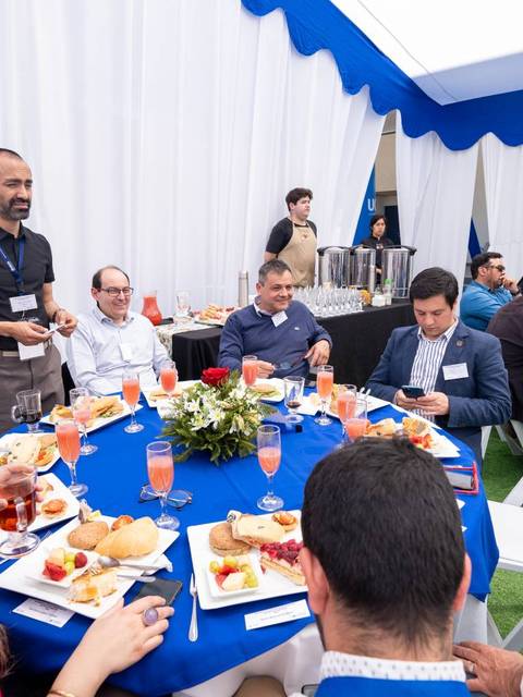 Un grupo de personas se reúne alrededor de una mesa decorada para un evento, disfrutando de comida y bebidas en un ambiente festivo.
