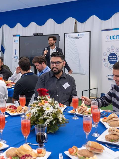 Un grupo de personas se encuentra en una mesa comiendo y escuchando una presentación en un evento.