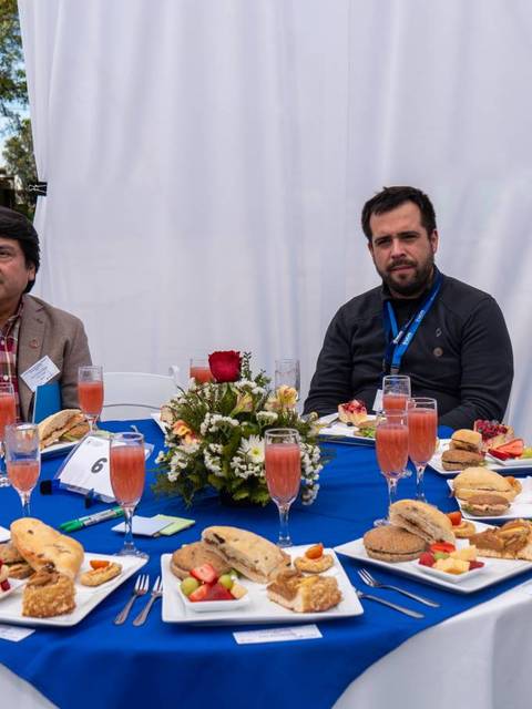 Grupo de personas sentadas alrededor de una mesa con comida y bebidas en un evento al aire libre.