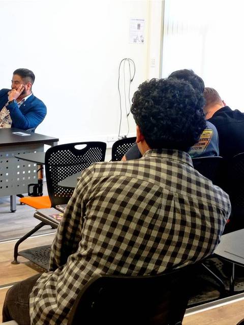 Una mesa de presentación con varias personas hablando y un grupo de espectadores sentados al fondo.