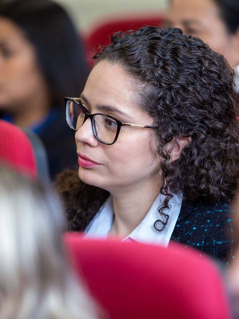 Una mujer con gafas escucha atentamente en un evento con otras personas al fondo.