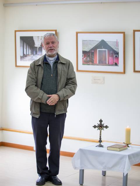 Un hombre de pie en una sala con fotografías en la pared y un altar con una vela y un crucifijo.