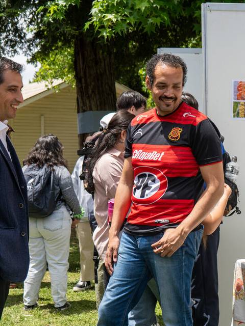 Un grupo de personas interactuando en un evento al aire libre, con un hombre alegre en camiseta de fútbol conversando con un conocido.
