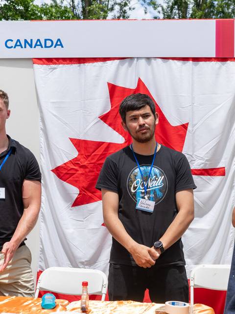Un grupo de personas conversando en un stand representativo de Canadá con una bandera en el fondo.