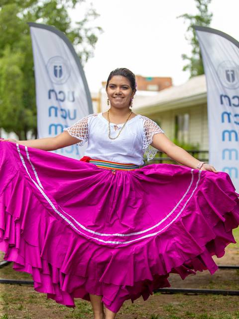 Una joven sonriente viste un traje tradicional con una falda larga de color rosa y se encuentra en un evento al aire libre.