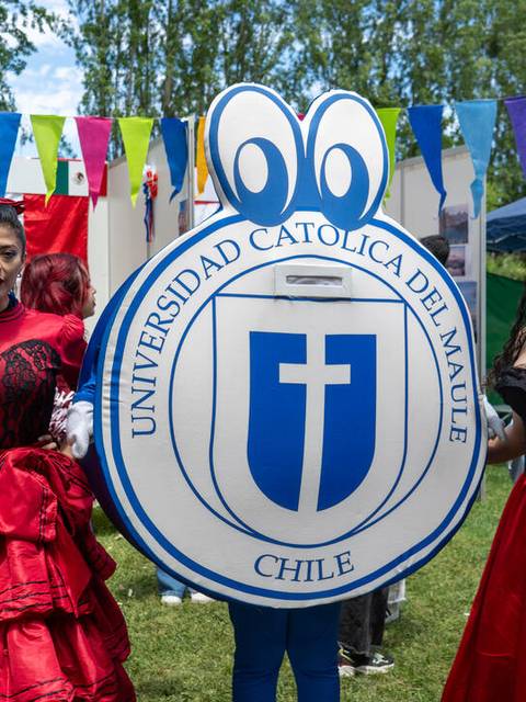 Dos mujeres vestidas con trajes tradicionales posan frente a un escudo de la Universidad Católica del Maule durante un evento cultural.
