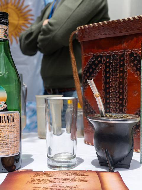 La imagen muestra una mesa con diferentes bebidas y un recipiente de mate, entre ellas un Fernet, una botella de Coca-Cola y un mate.