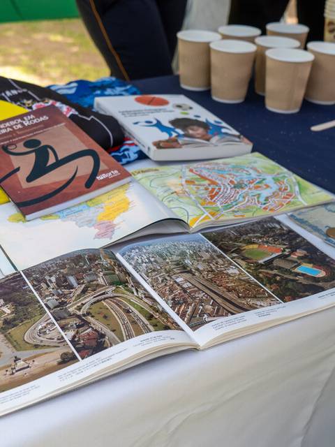 Una mesa con libros, mapas, camisetas y vasos de cartón en un evento al aire libre.