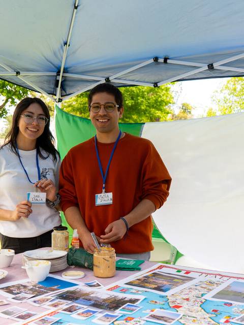 Dos jóvenes sonrientes se encuentran en un stand decorado con una bandera italiana y una mesa llena de fotos y alimentos.