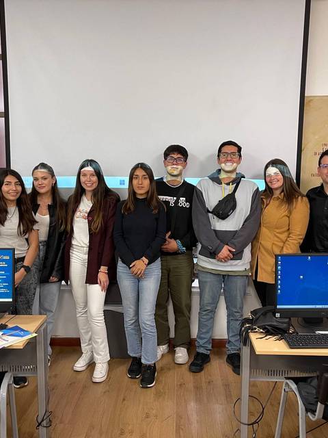 Grupo de estudiantes posando frente a una pantalla en un aula de computación.